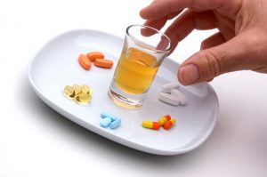 Hand reaching for a small glass of amber liquid, in the middle of a plate, surrounded by piles of pills.
