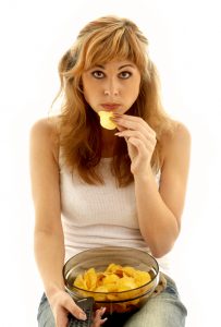 Young woman with blond highlights sits with a bowl of potato chips on her lap, lifting one to her mouth.