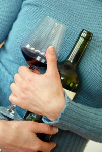 Torso of a woman wearing a blue sweater, hugging a bottle and glass of wine to her chest.