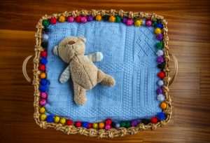 A cuddly teddy bear lying on a blue crochet cushion with colorful bobble edging, in a basket.
