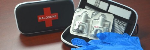 An open first aid kit showing the red cross on the cover, and in the open base, two narcan nasal sprays and blue nitrile gloves.