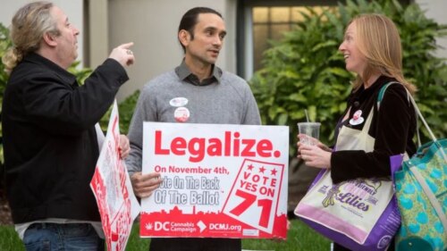 man holding 'legalize' sign stands between man making angry gesture and woman smiling agreement