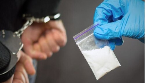 hands in handcuffs, background, foreground a blue nitrile-gloved hand holding a small baggie of white powder