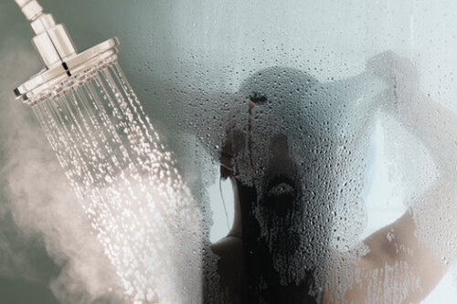 Showerhead at left on full with steam, in front of a steamed-up shower door with a person behind it.