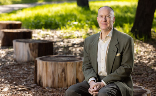 Mr. Humphreys in an outdoor setting, sitting on a section of tree trunk with hands folded in his lap, smiling pleasantly