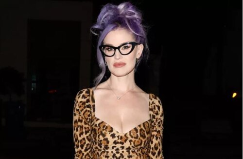 young woman (Kelly Osbourne) in low-neck leopard print dress, with purple hair and thickblack-rimmed cat-eye glasses.