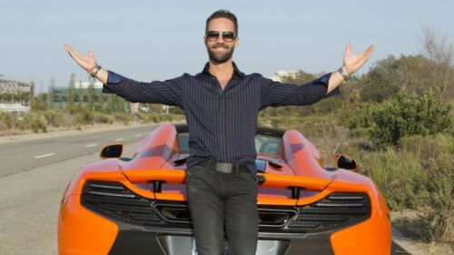 Bearded white guy in sunglasses standing in front of flashy orange convertible with arms spread, looking pleased with himself.