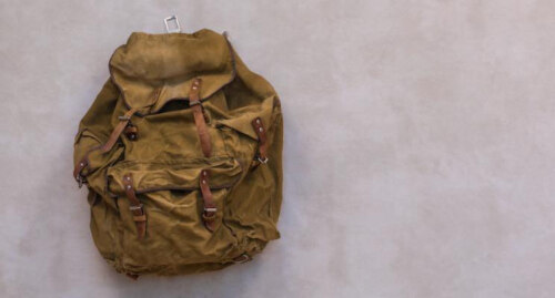 a brown knapsack with leather straps closing many pockets, off-center to the left against a gray background