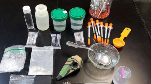 An array of syringes, small bags of powder, a pipe, spoons, bowls, bottles, etc., on a dark background.