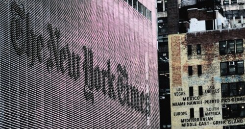 The frontage of the New York Times building with the name/logo angled from the left, and another urban building front on the right - predominantly tones of gray